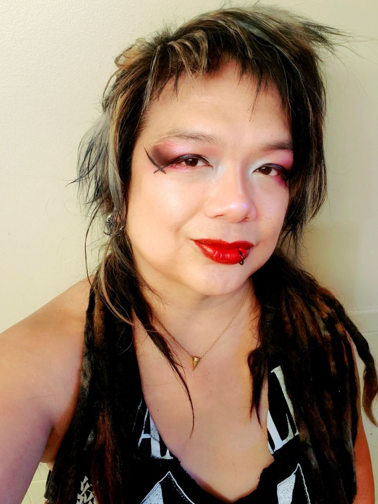 Kei chest up in a black with white print EVANGELION tee cut into a long halter tank, holster harness straps amd dreads forward on shoulders, red lipstain and white to pink and red with black dust outer eyeshadow and pink undershadow with crossed puppy eyeliner, bangs and wispys styled down and left messy, room vanity lighting.