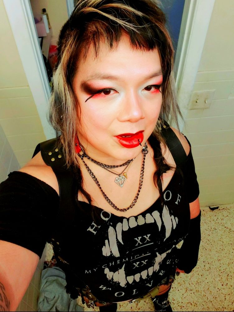 Downward angle selfie of Kei, a tan heavy set Filipunx Trans Woman, with right arm exiting bottom left of frame to hold up camera, black Chelsea boots with cutoff shorts and a black MCR HOUSE OF WOLVES tee cut into a deep wide scoopneck with black bracer straps over shoulders, short black dread tips forward on shoulders with a gunmetal and red strap slipchain collar, die-cut diamond and crass cone pendant necklaces, red lipcolor and white to red and black dust outer eyeshadow with red undershadow and extended puppy eyeliner, black and bleached side wispys down and choppy black bangs framing face, background is a white bathroom doorframe with light yellow walls, room vanity lighting.