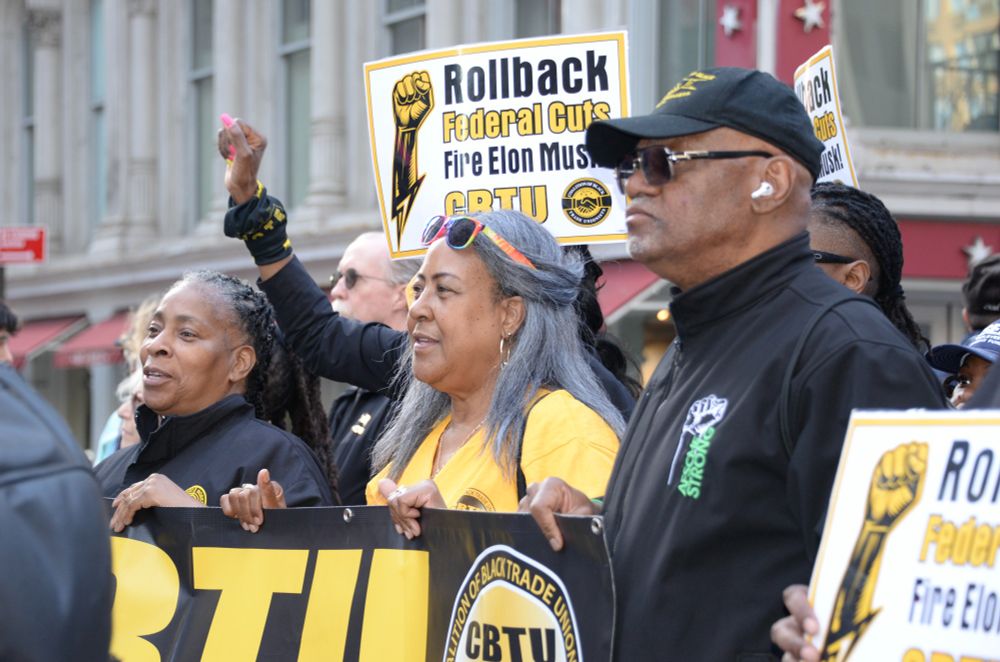 Workers dressed in black and yellow hold up signs that read, "Rollback Federal Cuts, Fire Elon Musk."
