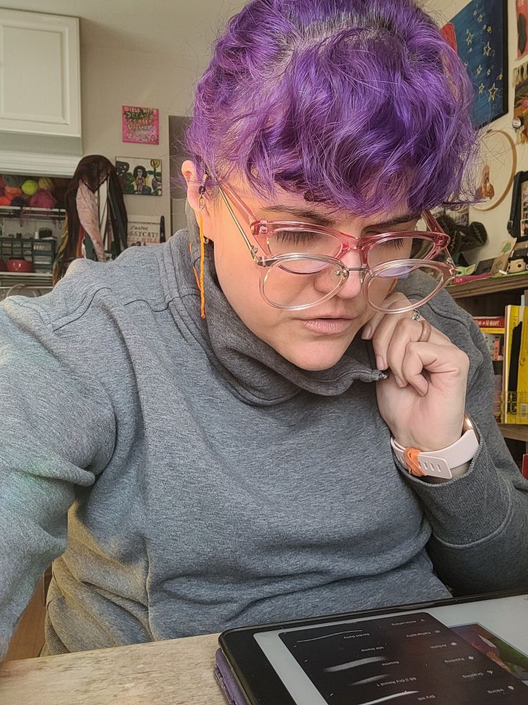 Morgan, an olive skin toned woman with purple hair, wears a pair of pink reader glasses under a pair of blue light glasses, staring at her ipad.