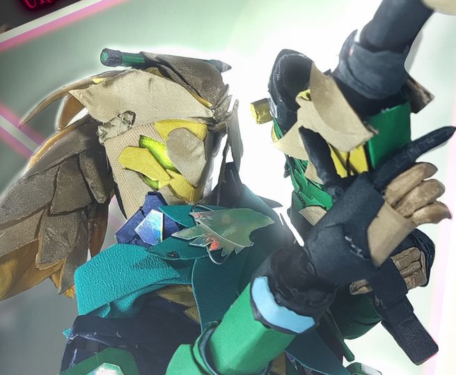 Shown is a photo of a handcraft of Goldbullet in his regular outfit, constructed of various paper and fabric materials. The photo shows his upper torso, and he is posed holding his sniper rifle, pointing it to the upper right of the viewer. 