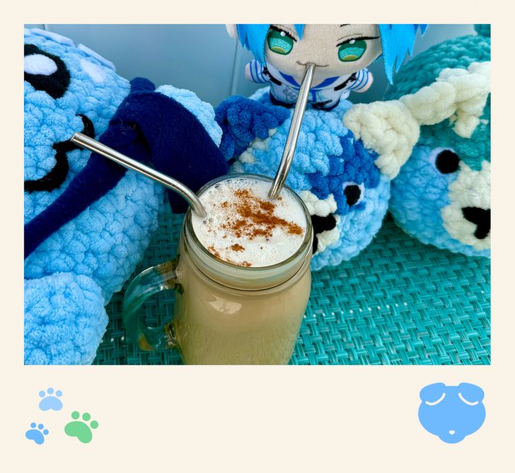 Altare and a plush Altventurer sit side by side, sipping from a glass filled with coffee, topped with foam and a sprinkle of cinnamon. Sitting alongside Altare and the Altventurer are two blue plush dogs.