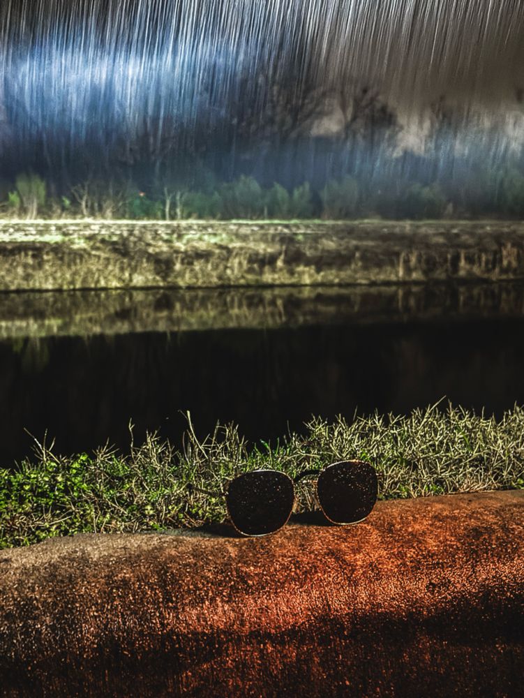 A pair of sunglasses lies on a rusty or reddish-brown curb, with a dramatic rainy landscape in the background. Heavy rainfall is visible as streaks against a dark, moody sky, and vegetation is reflected in what appears to be a body of water.