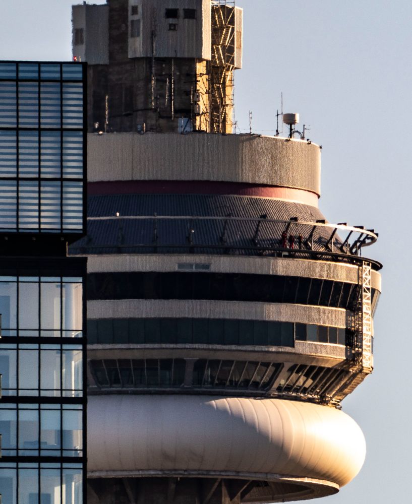 The CN Tower looms over an apartment building in the foreground, with little in the frame apart from the main pod of the tower. A little off-center is a small group of people walking around the edge of the CN Tower (the activity is appropriately called the "Edge Walk"). They have just rounded the north-facing side of the tower, now brightly lit by the setting sun.