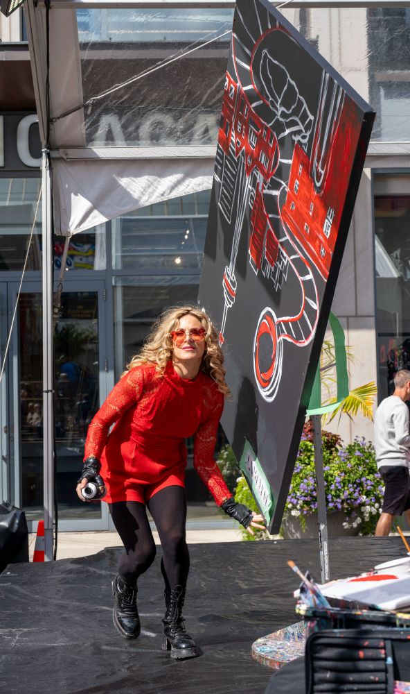 A Toronto artist, sunlit  on an open stage mid-day on a street in Yorkville, twirling a large painting she is in the middle of completing as she dances toward the photographer, smiling for the camera.