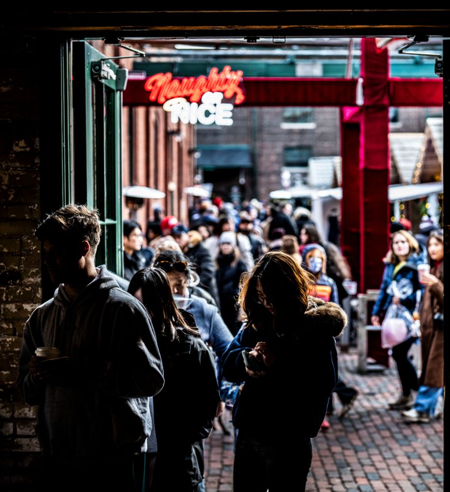 A tight view of a lineup at the entrance to a building, the lower center of the photo filled by a young woman, staring down at her hand and phone, brown hair backlit. The background beyond her is framed by a row of small cabins on one side, a red brick building on the other, filled with a crowd of people, all thrown out of focus separating and isolating her. Above it all is a neon sign "Naughty or Nice", which is a themed holiday bar at the Distillery Winter Village.