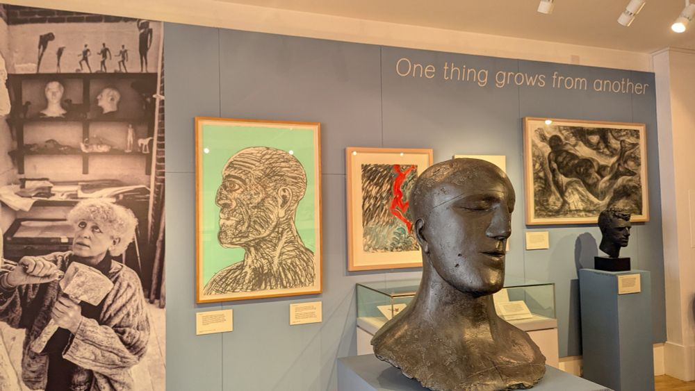 Background shows image of Elizabeth Frink with three prints. Front right is Tribute III - a large sculpted head. Background right, a smaller sculpted head.