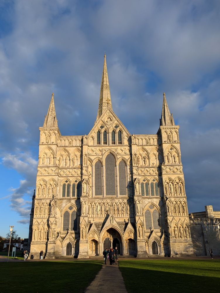 View of Salisbury Cathedral's west end bathed in late afternoon sunshine. Shadows cast across the grassy foreground. Background of blue sky with fluffy white grey clouds.