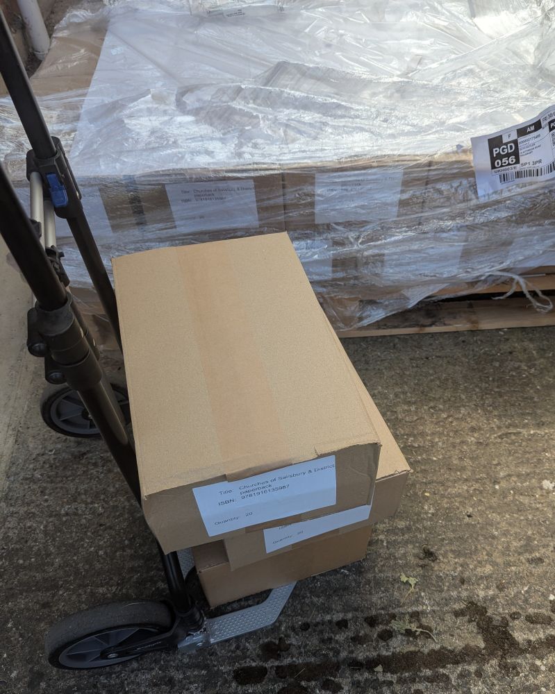 3 brown boxes on a sack trolley with a pile of boxes behind, stacked on a wooden pallet & covered in plastic.