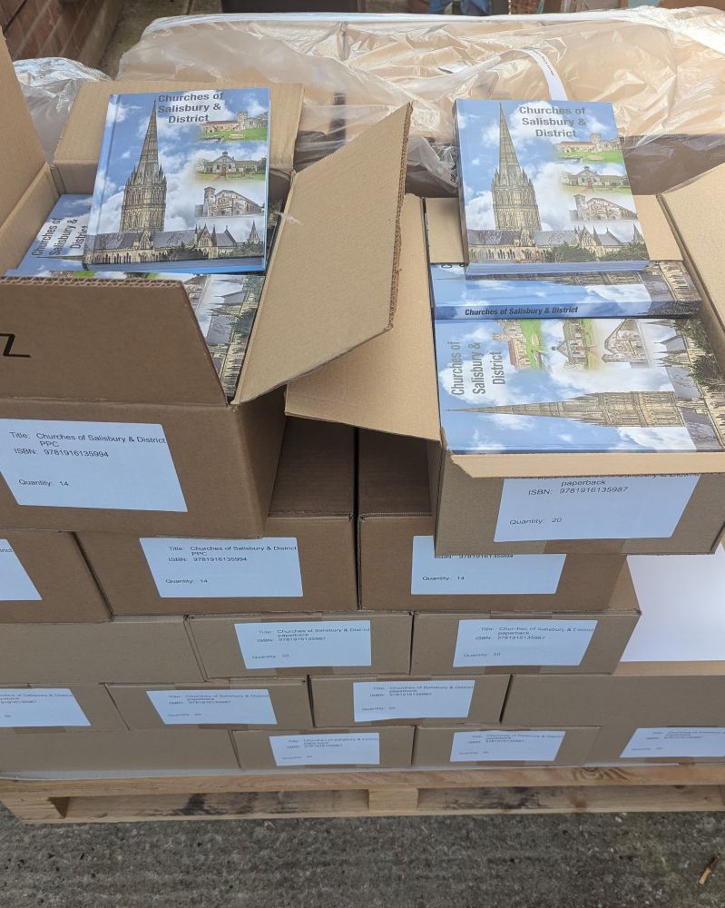 A pile of brown labelled boxes on a wooden pallet with two boxes open to show books titled Churches of Salisbury & District. The book cover shows Salisbury Cathedral & 3 other churches.