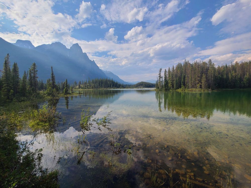 A calm, crystal clear lake reflects the surrounding forest and cloudy blue sky. Sharp, tall mountains can be seen in the upper left corner.