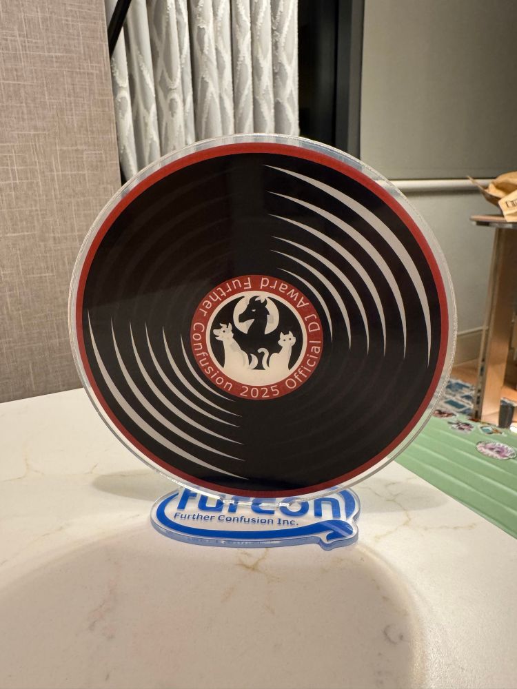 An acrylic vinyl record about 6 inches tall, with the Further Confusion logo in the middle surrounded by the text "Further Confusion 2025 Official DJ Award". The base is also acrylic and says "Furcon" in the style of the Intel logo. It's a major award!!