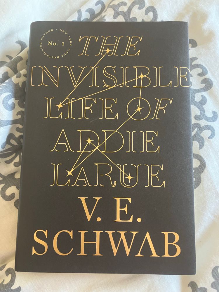 A book, “The Invisible Life of Addie LaRue by V.E. Schwab” sitting on a floral bedsheet.
