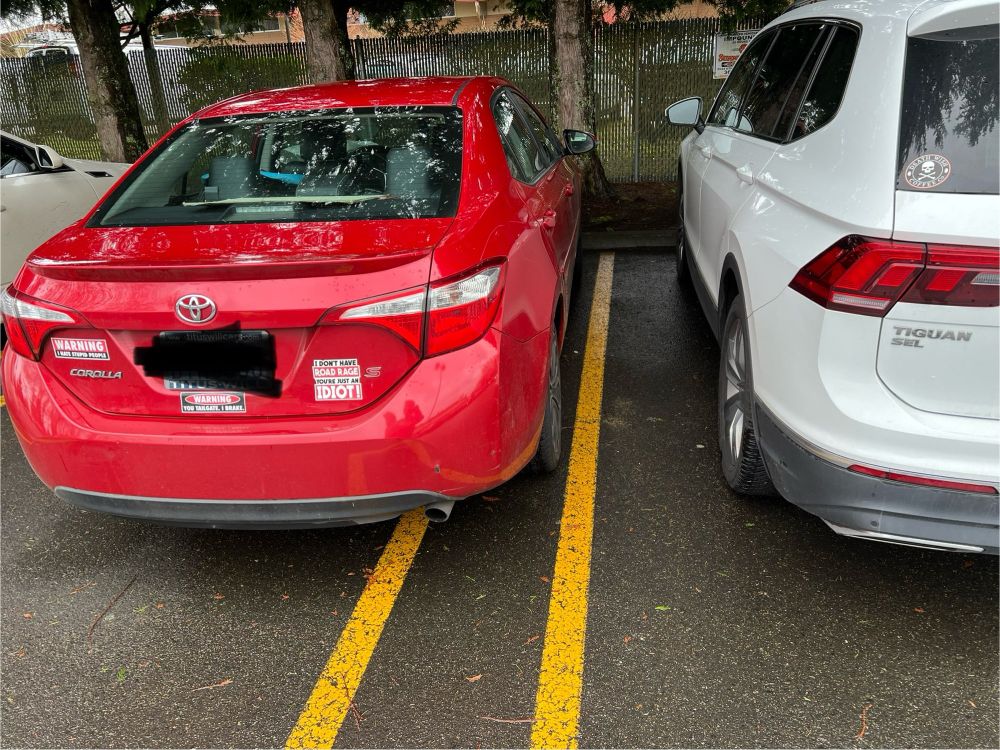 Two cars parked in a lot. One white car is parked neatly in the parking space. The second red car is parked to the left of the first, wildly over the line. Three bumper stickers are visible on the red car, reading "WARNING: I HATE STUPID PEOPLE", "WARNING: YOU TAILGATE, I BRAKE", and "I DON'T HAVE ROAD RAGE, YOU'RE JUST AN IDIOT"