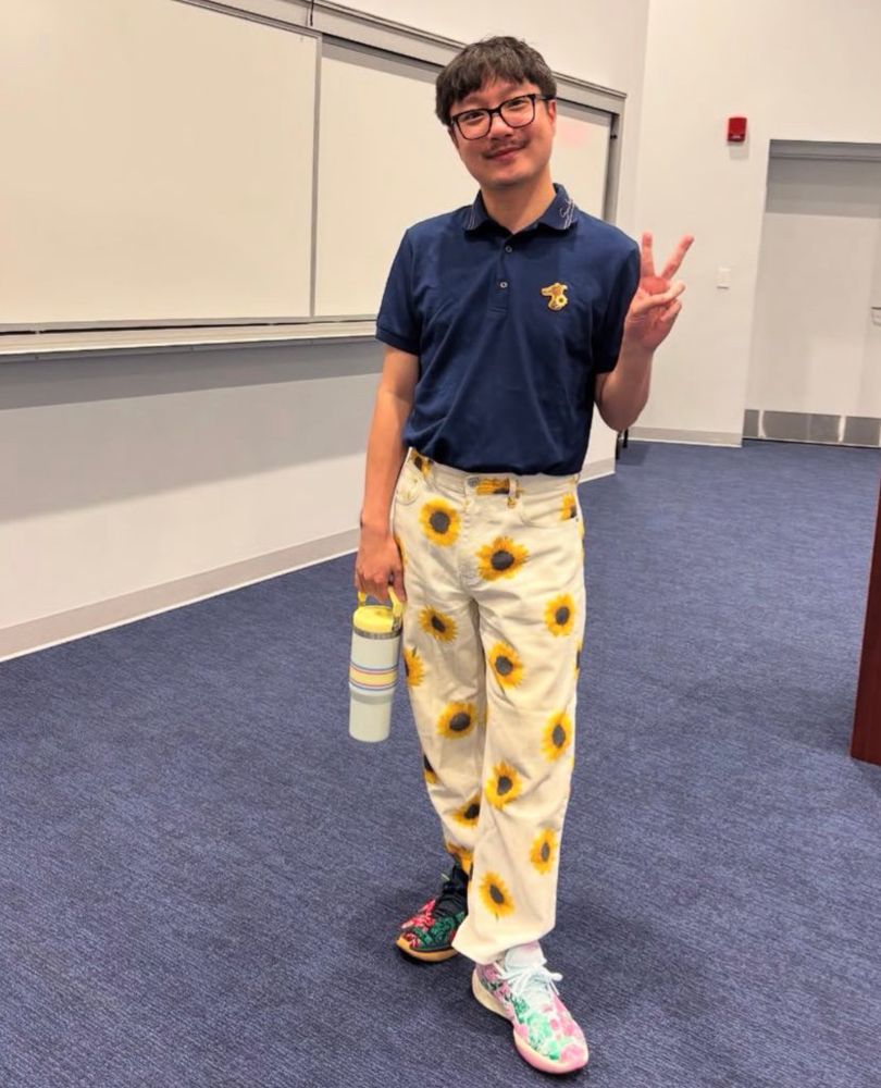 i’m standing in an auditorium & i’m wearing black glasses, a blue shirt with a dog logo on the chest, white trousers with a sunflower print, & two different sneakers with complementary embroidered floral patterns