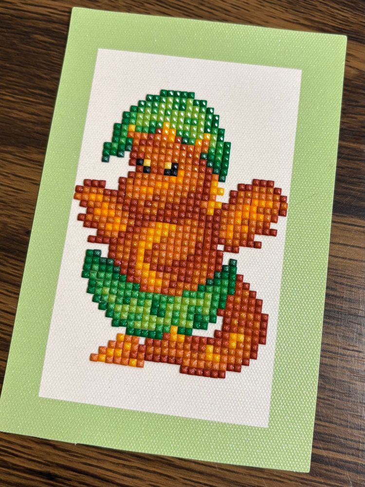 Small diamond painting of a pixelated, orange-brown dinosaur who just hatched. He has green shell fragments on his head and feet, and is on a white cardstock background, framed by a light green cardstock.