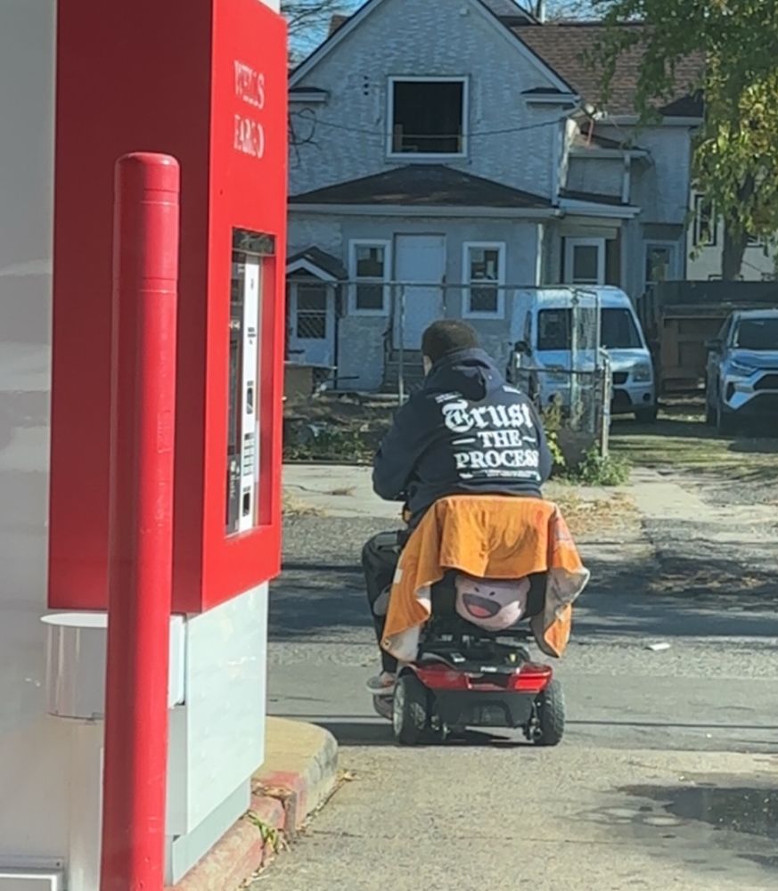 A man scootering away from a WellsFargo ATM wearing a shirt that says Trust the Process