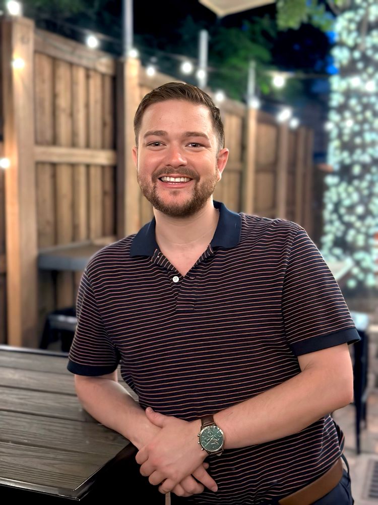 I am leaning against the table, smiling in a dark blue polo with orange stripes and a green fossil wash with a brown leather band