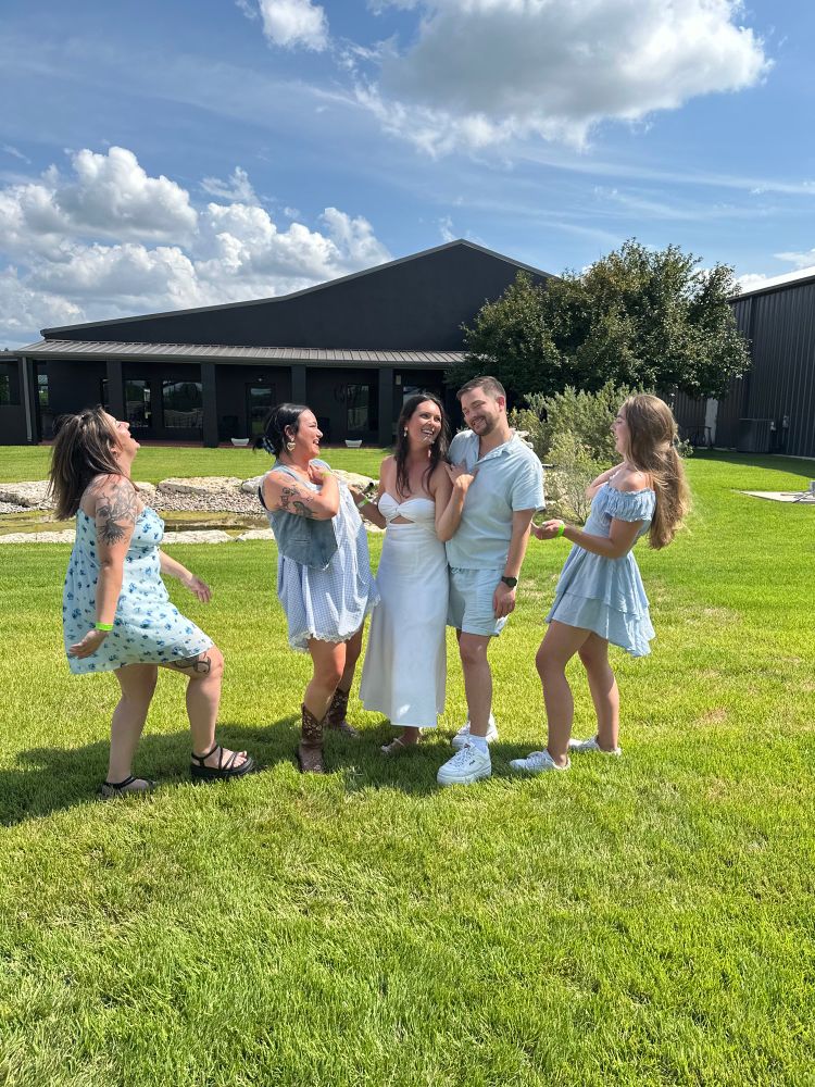 A woman and a white dress stands in the middle of a field, flanked by four friends two on each side and light blue clothing. It’s an action shot. Where three friends are laughing. One is making a goofy smile. And the bride to be in White is smiling gracefully