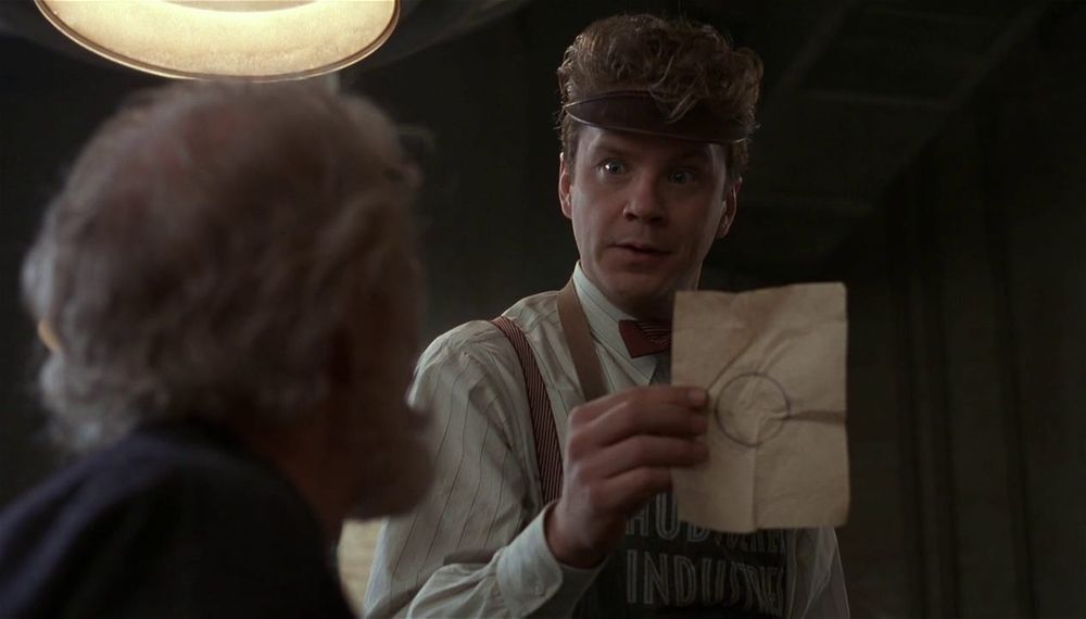 Still frame from The Hudsucker Proxy. Tim Robbins’ character holds up a wrinkled piece of paper with a circle drawn in pencil in the center of the page.