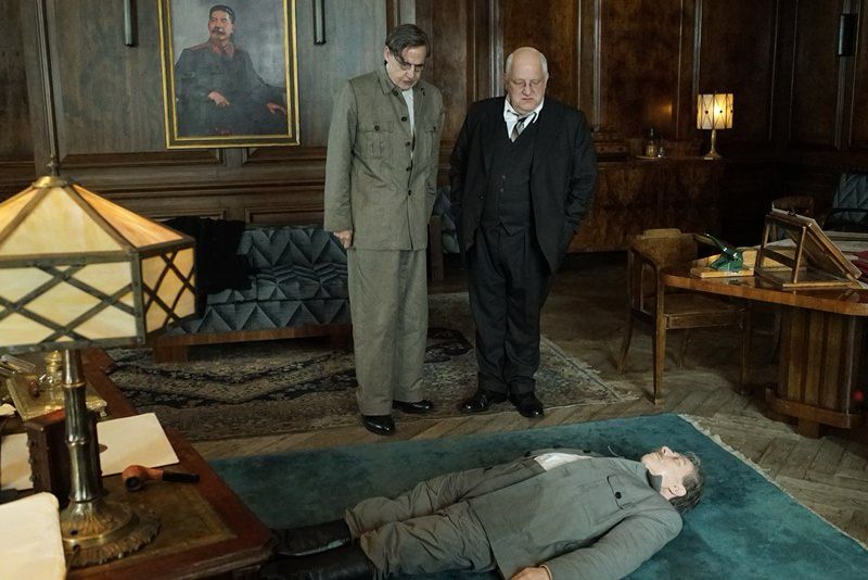 Screenshot from The Death of Stalin. Josef Stalin lies dead on the floor of his office while two officials stand next to him, looking down at the body. 
