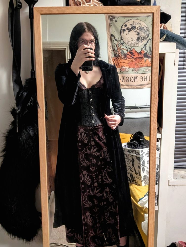 Trans person in a purple velvet dress, corset, and black velvet trench coat