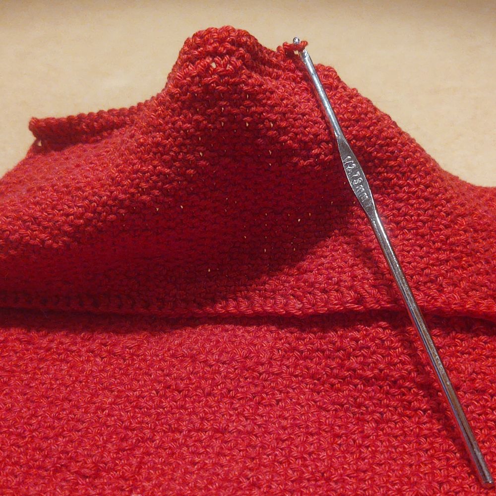 Red knit fabric, with crocheted seams and a silver crochet hook working in the far edge.