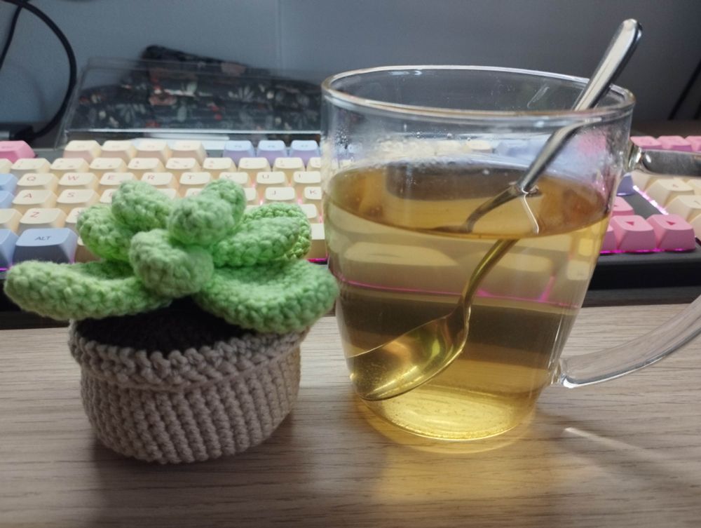 Imagen de una taza de cristal trasparente con una infusión de color anaranjada. A su izquierda, una plantita de crochet. De fondo un teclado de ordenador en colores amarillo, morado y rosa.
