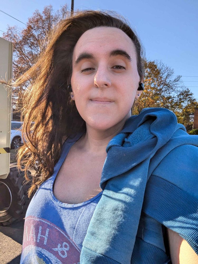 Stella, looking at the camera. She is sleepy and the wind is blowing her hair. She's wearing a blue tank top with a teal-ish sweatshirt over her shoulder.