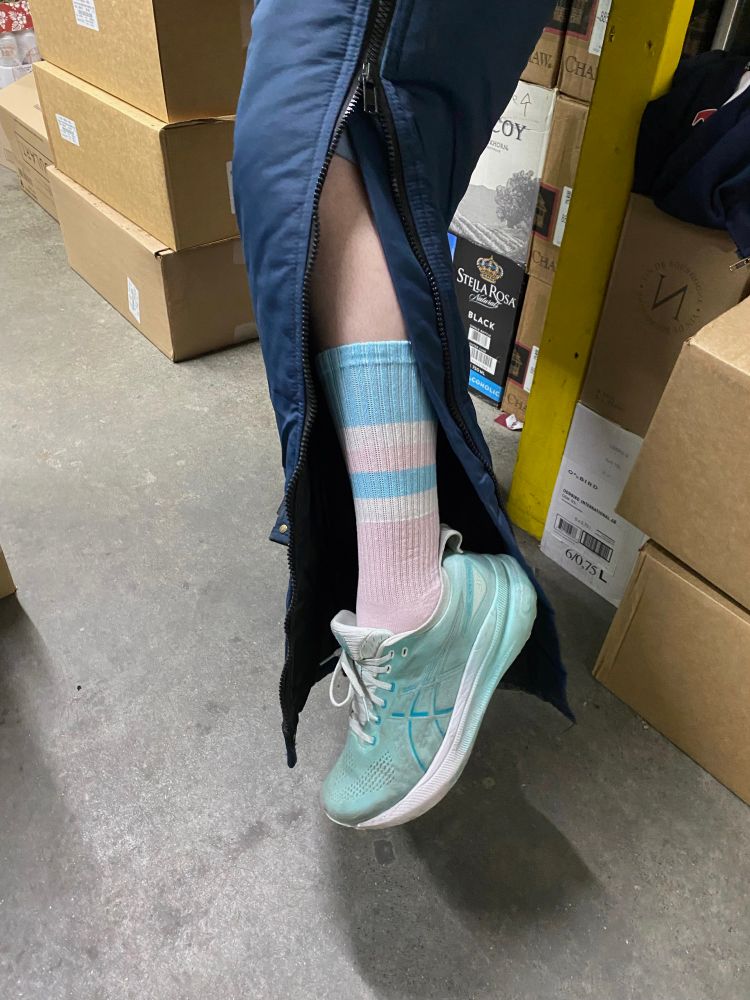 close up of white person’s leg wearing a snow suit with the zipper undone to reveal a knee sock in the colors of the trans pride flag 