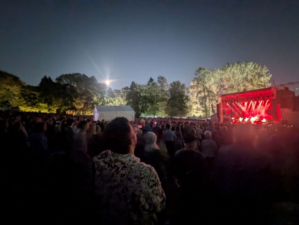 A red lit sage on the right, a large audience in a dark park, a bright moon rising above trees in the center.