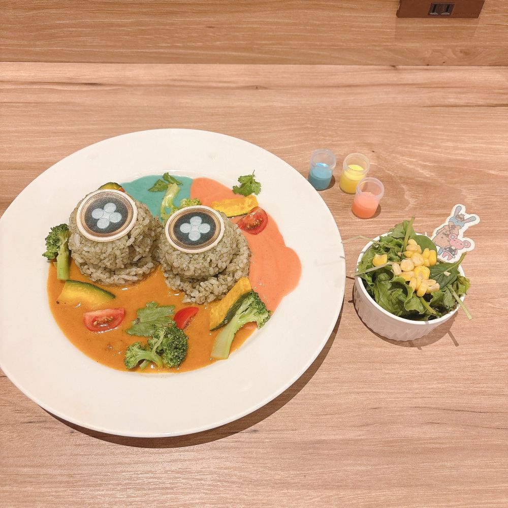 カプコンカフェのホームズの試験管カレーの写真。ホームズの試験管に入っているカラフルな試薬のような青、ピンク、オレンジ色の3色のカレールーを敷き詰めた上に、ホームズの頭に載っているメガネ？を模したご飯がのっている。
右横には小さいサラダとこれまた3色のドレッシングが添えてある。