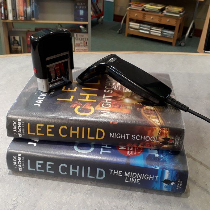 Two Jack Reacher books by Lee Child on a library desk beside a date stamp and a handheld scanner