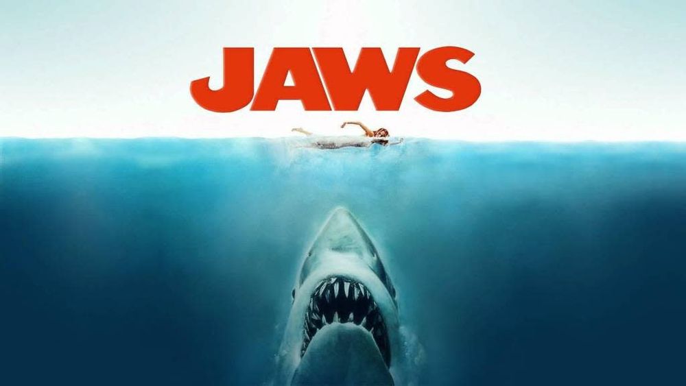 A poster for the movie Jaws with the shark swimming up under a woman swimming on the surface. The text Jaws is at the top.
