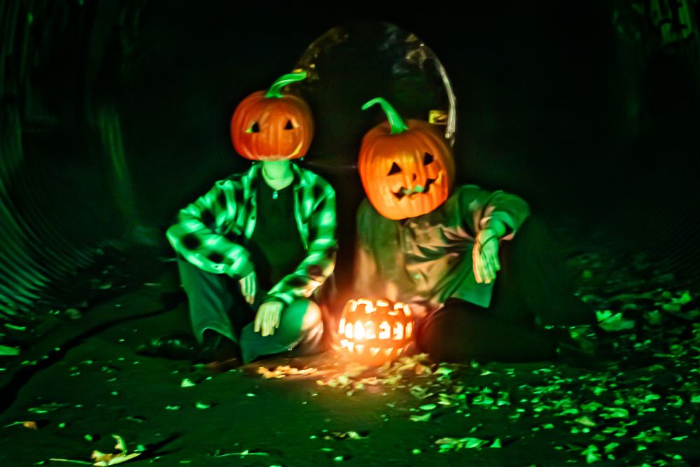 Two people wearing comfy street clothes and pumpkins on their heads sit in a tunnel lit by a green light. There is a pumpkin between them with a fire going in it. 