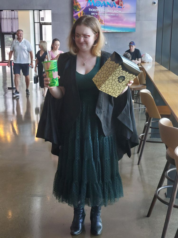 a young woman with blonde hair wearing green and a cape, holding merch for the movie "wicked" and looking at it quizzically