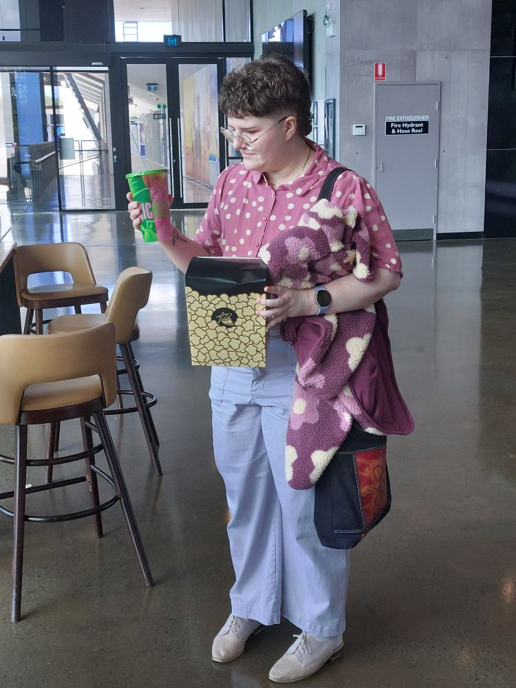 a nonbinary person with short brown hair wearing pink, holding merch for the movie "wicked" and looking at it quizzically