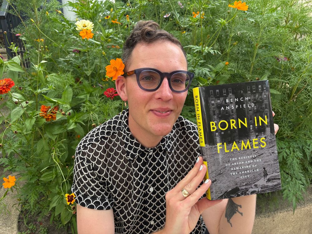 I am sitting in front of flower garden holding a copy of my new book, BORN IN FLAMES.