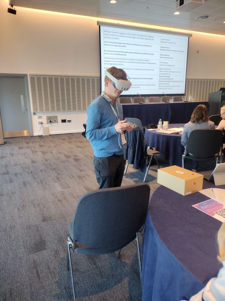 Prof Ed Hutchinson stands in a seminar room wearing a VR headset and holding VR controllers