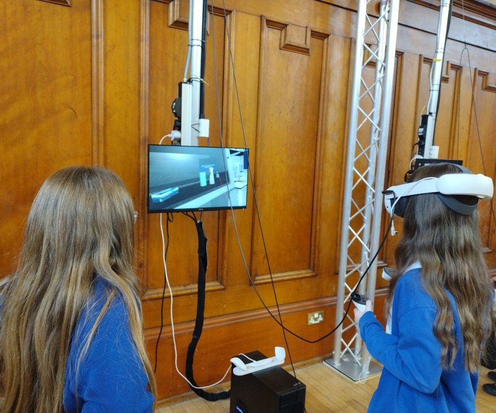 Two pupils in blue school jumpers stand in front of a suspended computer monitor showing a microbiological safety cabinet in virtual reality. One of the pupils wears a headset and holds a controller in each hand while the other watches what they are doing on the screen 