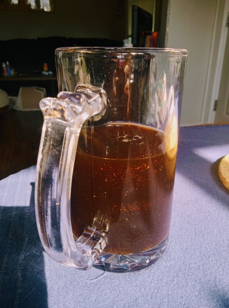 A photo of a glass cup of cold coffee resting on a table, with a living area in the background. The coffee has small shiny glitter specks floating throughout. 