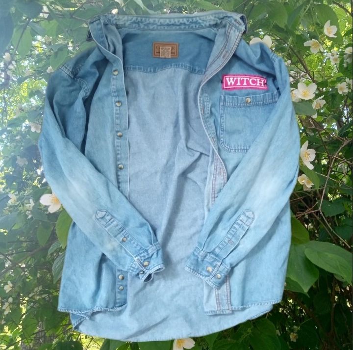 A light-wash denim jacket with an embroidered pink nametag that reads "witch," rests above a background of leaves and white flowers. 