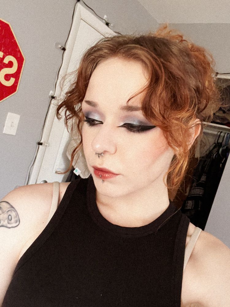 A feminine, red haired person with muted, but dramatic makeup and big eyeliner looking down towards the floor