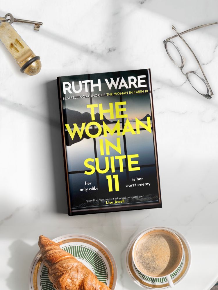 Flat lay of a copy of The Woman in Suite 11 by Ruth Ware on a marble background, surrounded by a croissant, a cup of coffee, and a hotel keychain with the number 11 on the fob.