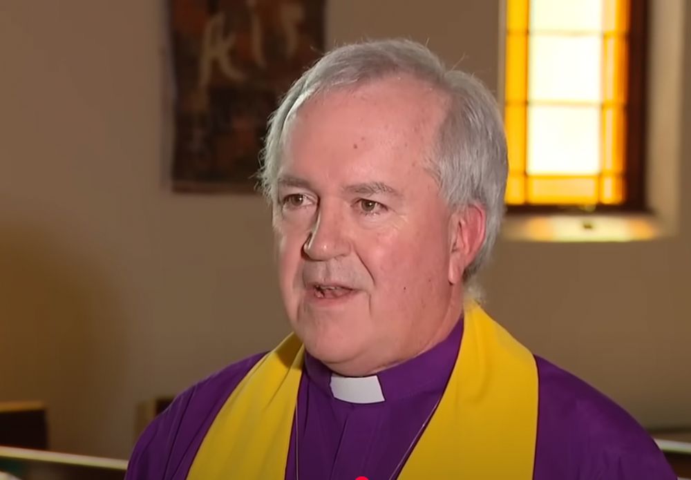 Rev. Benedict Atherton-Zeman, in a purple clerical shirt with goldenrod stole (which says Side With Love and has heart logos), in a still from his interview inside the sanctuary of his UU congregation in St. Pete.