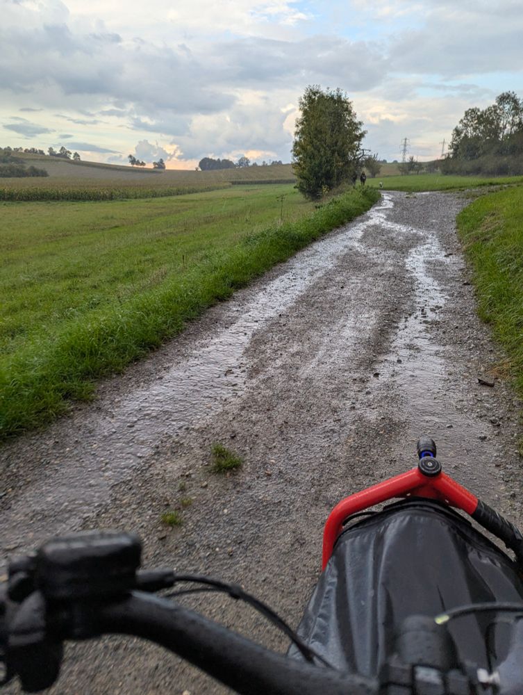 Ein Lastenrad im Vordergrund, auf einem sehr nassen und matschigen Feldweg. Im Hintergrund dramatische Wolken 