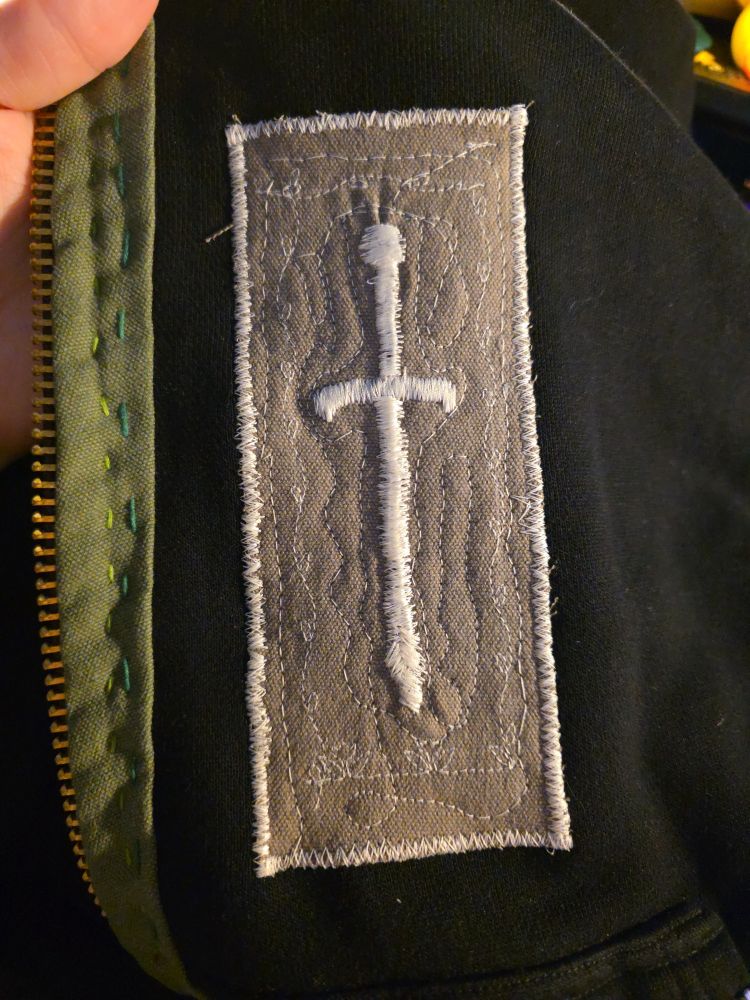 A beige patch with a messily machine embroidered sword in white thread