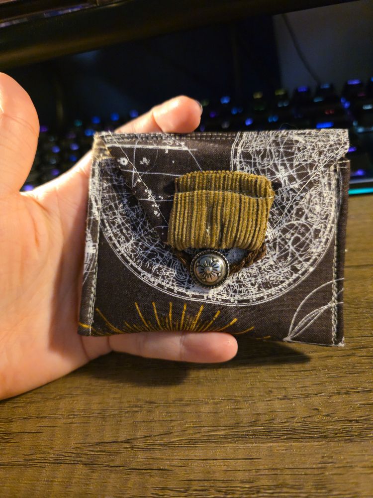 A brown envelope shaped fabric pouch with celestial themed pattern, a flower button, string closure with a brown corduroy square holding the string in place