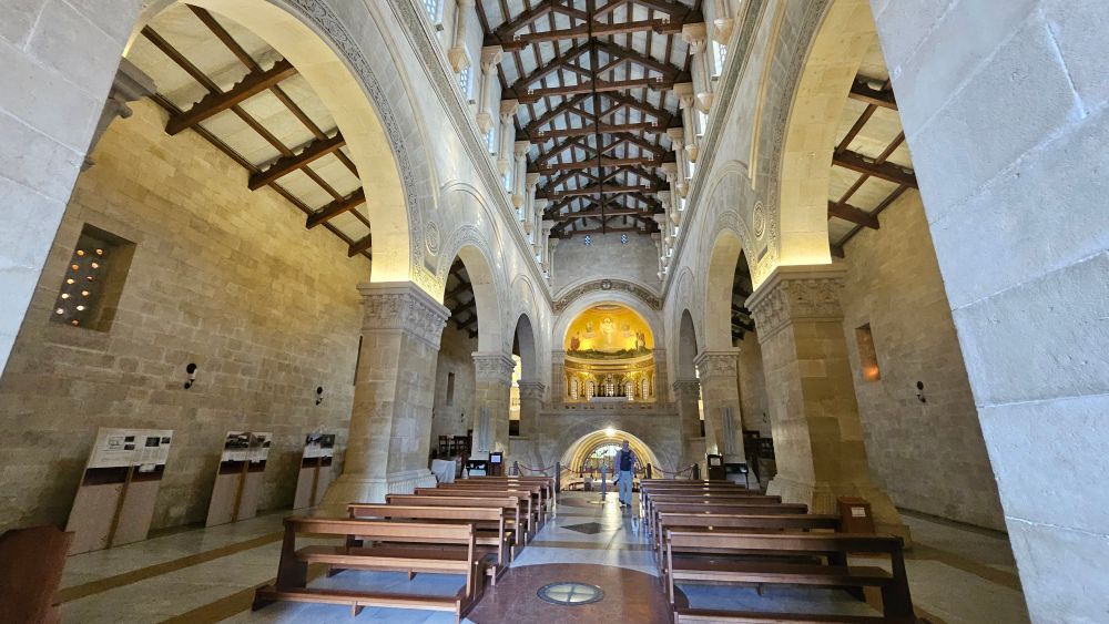 The inside of a cathedral with stone walls and wooden accent beams on the roof. Pews made of wood are also seen
