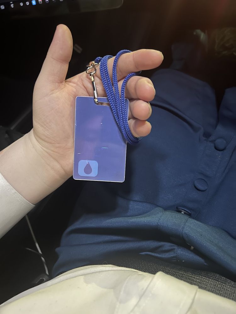 a hand holding up a Lumon MDR badge, blue with raindrop logo, camera is looking down, photographer is wearing a blue skirt and white button up shirt